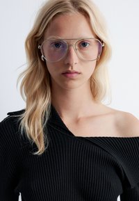 Silberne Metallbrille mit großen, klaren Gläsern. Das Gestell hat eine runde Form. Die Person trägt ein schwarzes, geripptes Oberteil mit einem trägerlosen Design.