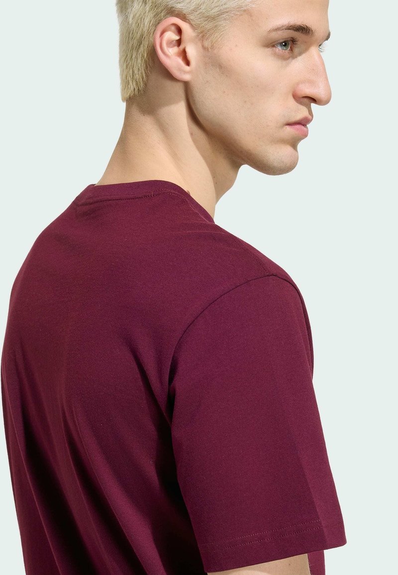 Jeune homme aux cheveux blonds courts portant un t-shirt bordeaux uni à manches courtes, vu de profil et de dos sur un fond clair.