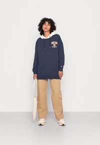 Námornícky oversized mikina s bielou kapucňou, s logom "Tommy Jeans". V kombinácii s béžovými cargo nohavicami a bielo-oranžovými teniskami; drží tašku.