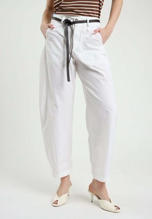 Donna con pantaloni bianchi a vita alta con cintura nera, mani nelle tasche, top marrone a righe e scarpe bianche aperte sulla punta con tacco su sfondo semplice.