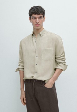 Ragazzo con capelli corti e ricci che indossa una camicia beige con bottoni e maniche rimboccate e pantaloni marrone scuro, in piedi davanti a uno sfondo semplice.