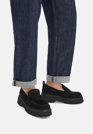 Osoba nosící tmavě modré ohrnuté džíny a černé semišové masivní platformové mokasíny na bílém pozadí.