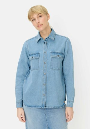 Blonde woman wears light blue denim jacket with buttoned chest pockets and matching denim skirt against plain white background.