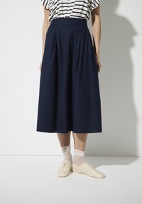 Marineblauwe geplooide midi-rok van lichtgewicht stof, met een hoge taille, gecombineerd met lichte schoenen en doorschijnende enkelkousen.