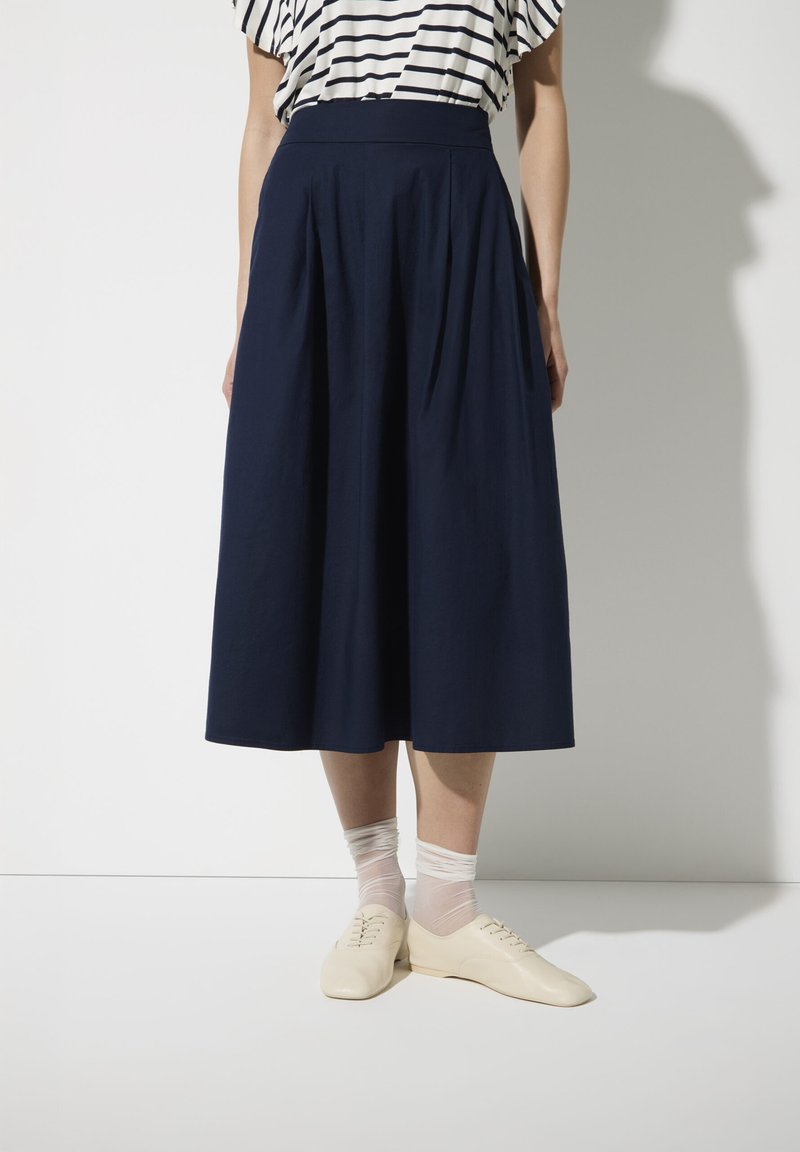 Marineblauwe geplooide midi-rok van lichtgewicht stof, met een hoge taille, gecombineerd met lichte schoenen en doorschijnende enkelkousen.