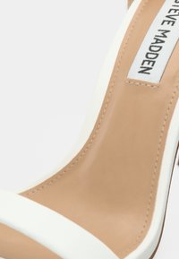 Λευκό και μπεζ πέδιλο με ψηλό τακούνι, ανοιχτή μύτη, φαρδιά λωρίδα πάνω από το πόδι και λείο, υφασμάτινο υλικό. Ορατό το λογότυπο της μάρκας.