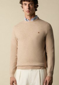 Maglione beige lavorato a maglia con scollo rotondo e maniche lunghe, caratterizzato da un logo discreto sul petto, indossato sopra una camicia a righe blu con colletto.