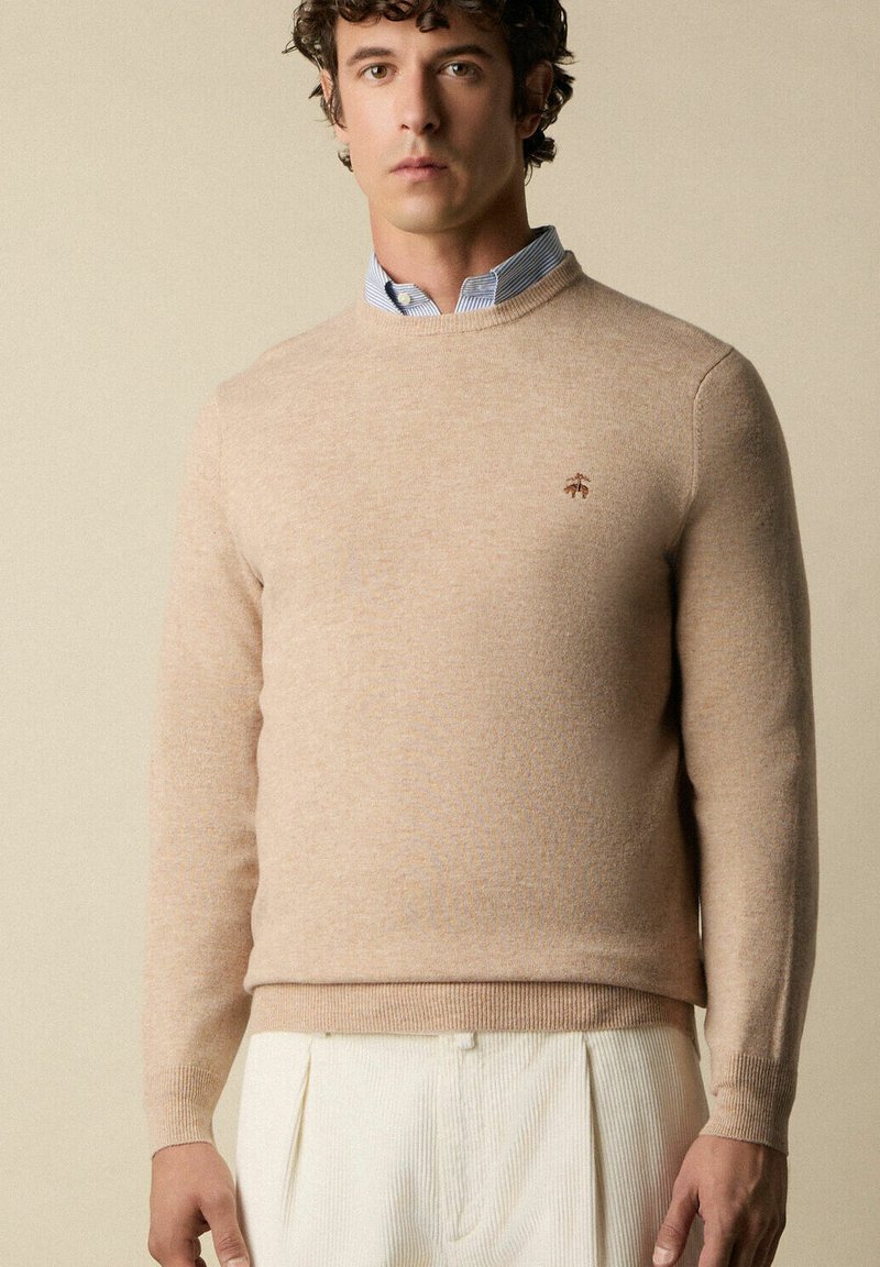 Maglione beige lavorato a maglia con scollo rotondo e maniche lunghe, caratterizzato da un logo discreto sul petto, indossato sopra una camicia a righe blu con colletto.