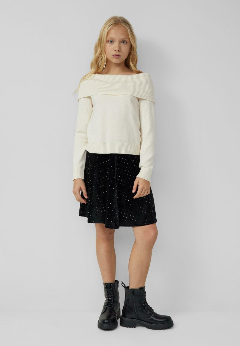 Crème off-the-shoulder trui, zwarte fluwelen rok met witte stippen, en zwarte veterschoenen met dikke zolen. Minimalistisch ontwerp.