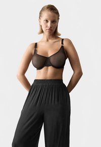 Sort gennemsigtig bh med bøjle, justerbare stropper og hægteslukning. Kombineret med sorte, løstsiddende bukser med elastik i taljen.