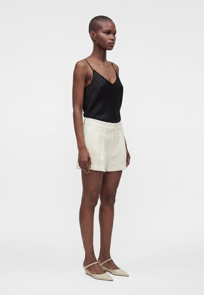 Femme debout de profil, portant un débardeur noir, un short blanc et des ballerines pointues couleur crème sur un fond blanc uni.