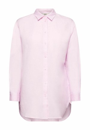 Kvinde iført en lyserød åben skjorte over en gul top, blå lige jeans og hvide sneakers, der står mod en hvid baggrund.