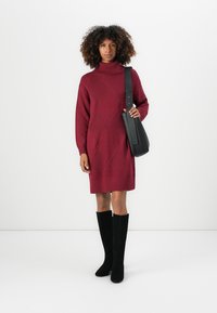 Bordeauxrotes Strickkleid mit hohem Ausschnitt und rautenförmigem Rippmuster. Es wird mit einer schwarzen Schultertasche und schwarzen kniehohen Stiefeln kombiniert.