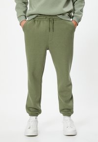 Pantalones de chándal verde oliva hechos de tela suave, con una cintura elástica y cordón ajustable, bolsillos laterales y puños de canalé en los tobillos.