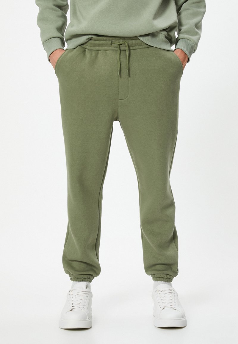 Pantalones de chándal verde oliva hechos de tela suave, con una cintura elástica y cordón ajustable, bolsillos laterales y puños de canalé en los tobillos.