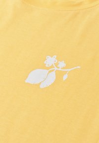 Gelber Stoff mit einer kleinen weißen Grafik von zwei Blumen und zwei Blättern, verbunden durch einen Stiel im Brustbereich.