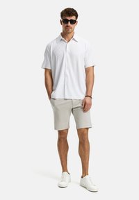 Korte mouwen, wit gestreept overhemd met knopen, lichtgrijze sweatshorts, witte sneakers en zwarte zonnebril. Eenvoudige, casual outfit.