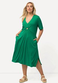 Vihreä mekko, joka on valmistettu sileästä kankaasta, syvällä V-pääntiellä, lyhyillä hihoilla, vyötäröllä solmittu yksityiskohta, sivutaskut ja halkiohelma.