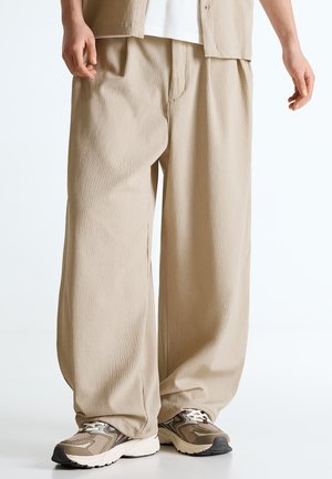 Beżowe, szerokie spodnie o fakturowanej strukturze, noszone z białymi trampkami i białą bluzką, pokazywane od połowy uda w dół, z rękami swobodnie opuszczonymi wzdłuż ciała.