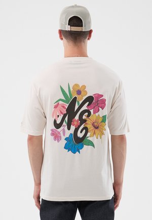 Hombre con camiseta blanca con diseño floral colorido y grandes letras "NE" en la parte trasera, combinada con una gorra de malla beige.