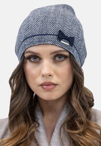 Gorro de punto azul y blanco con un patrón que presenta un lazo decorativo en navy y un acento plateado. Textura suave, diseño de parte superior redondeada.