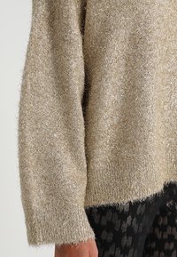 Beżowy puszysty sweter z metalicznymi nitkami, okrągłym dekoltem, luźnym krojem i ściągaczem. Teksturowana tkanina o miękkim, ciepłym wyglądzie.