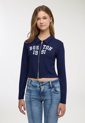Giacca corta con zip blu navy a trama a coste, con la scritta "BOSTON 1991" in bianco sul davanti. Abbinata a jeans azzurri.