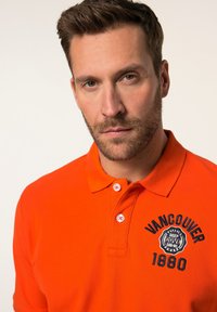 Oranžové polo tričko s límečkem, se třemi knoflíky a vyšitým logem "Vancouver 1880". Vyrobeno z texturované látky.