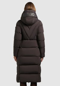 Puffjacka i mörkbrunt, med en lång design, oversize huva och KHUJO-logotyp på baksidan av kragen. Texturerad, kviltad tyg med synliga sömmar.