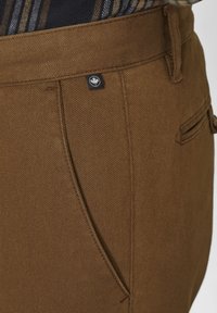 Brown textured trousers with a straight fit, featuring front pockets and a small brand tag near the waistband.