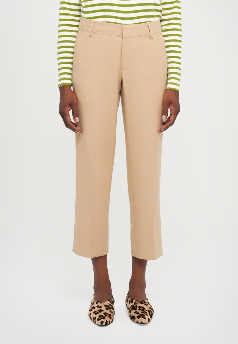 Personne portant un pantalon beige coupé, un haut à manches longues rayé vert et blanc, et des chaussures slip-on à motif léopard, debout devant un fond uni.