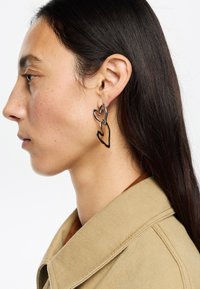 Pendientes en forma de corazón en negro y dorado con un diseño abierto en capas. Llevados por un modelo, mostrando una textura metálica suave y una forma elegante.