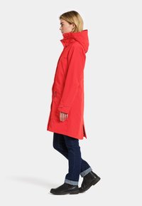 Nainen tummissa farkuissa ja mustissa saappaissa, pukeutunut polvipituiseen punaiseen hupulliseen takkiin, seisoo sivuprofiilissa yksinkertaista taustaa vasten.