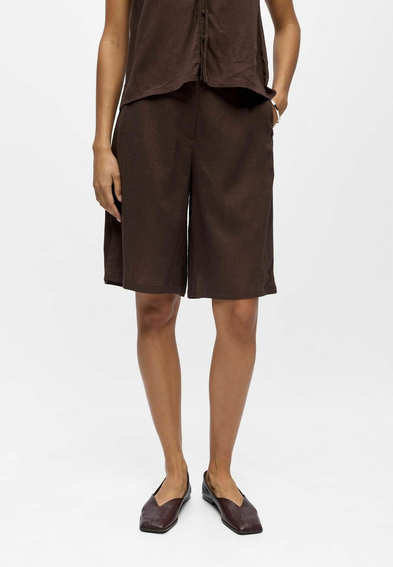 Personne portant un short marron ample jusqu'aux genoux, un haut sans manches marron assorti et des ballerines pointues brun foncé, debout avec une main dans la poche.
