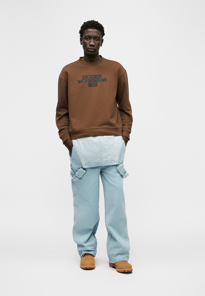 Man wearing brown sweatshirt with "Dickies Workwear 1922" text, light blue carpenter jeans with straps, and tan work boots, standing with hands in pockets.