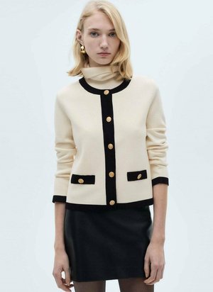 Beige sweater with black accents, gold buttons, and a mock neck design. Features two front pockets and three-quarter sleeves. Black skirt below.