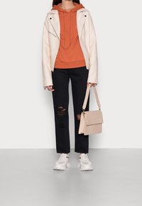 Person som bär en krämfärgad motorcykeljacka, orange huvtröja, svarta trasiga jeans, vita sneakers och håller en beige axelväska framför en enfärgad bakgrund.