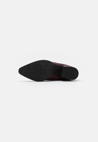 Chaussure bordeaux avec un dessus texturé, présentant un design à bout pointu, une semelle noire plate et un embossage décoratif sur la semelle extérieure.