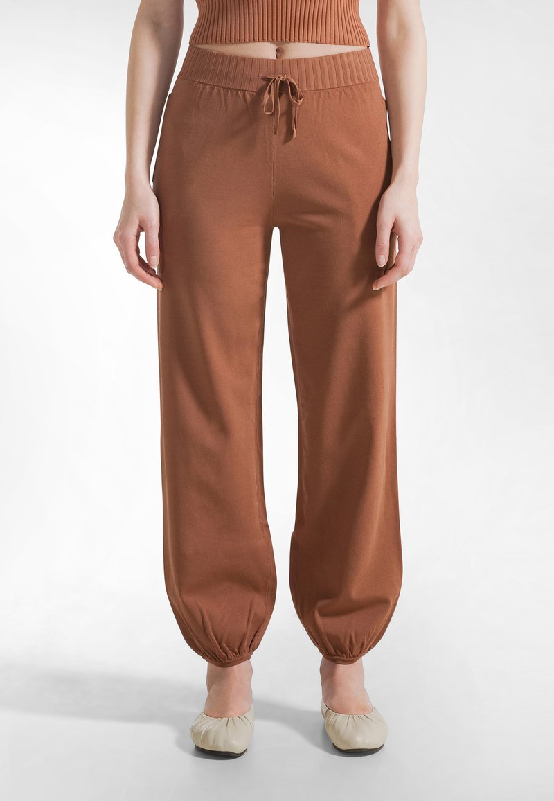 Persona con pantalones marrones de tiro alto con cordón y puños elásticos combinados con zapatos planos beige, de pie sobre un fondo blanco.