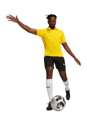 Geel polo-shirt, zwarte korte broek en witte sokken met zwarte accenten. Speler is in beweging met een voetbal.
