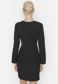 Femme aux cheveux blonds en chignon, portant une robe noire ajustée à manches longues, se tenant de dos contre un fond blanc uni.