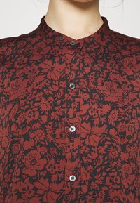 Červená halenka s květinovým vzorem na černém pozadí, se stojáčkem a zapínáním na knoflíky. Vyrobena z lehkého materiálu se hladkou texturou.