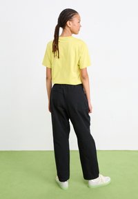 Gelbes kurzärmliges T-Shirt, kombiniert mit lockeren schwarzen Hosen und weißen Sneakern mit grünen Sohlen. Modell von hinten gezeigt.