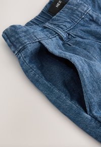 Mėlyni džinsiniai šortai su tekstūruota apdaila. Turi juosmens juostą su kilpelėmis ir šonines kišenes, atspindinčias kasdienišką dizainą.