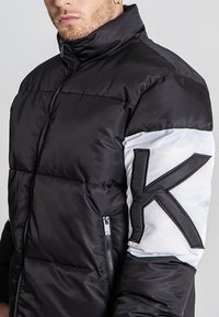 Chaqueta acolchada negra con un cuello alto, presenta una manga blanca con un gran logotipo negro 'K' y un bolsillo con cremallera en el lateral.