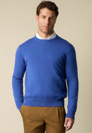 Maglione blu a maglia con scollatura rotonda, maniche lunghe e polsini e orlo a coste. Indossato sopra una camicia bianca con colletto.