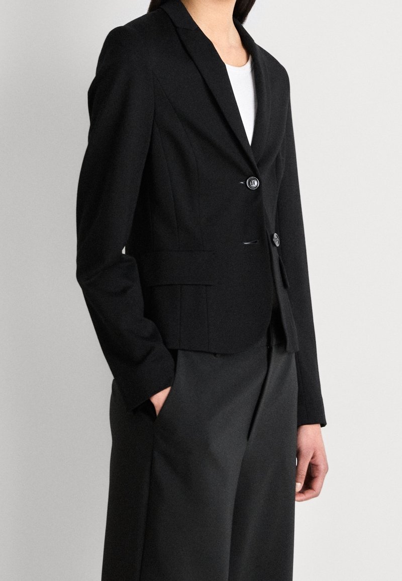 Femme portant un blazer noir ajusté sur un haut blanc avec un pantalon noir à jambes larges, une main dans la poche, sur un fond clair uni.