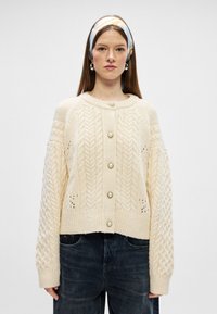 Cárdigan de punto crema con patrones de trenzado, escote redondo y botones decorativos de perla. Presenta mangas largas y texturizadas, y un diseño corto.