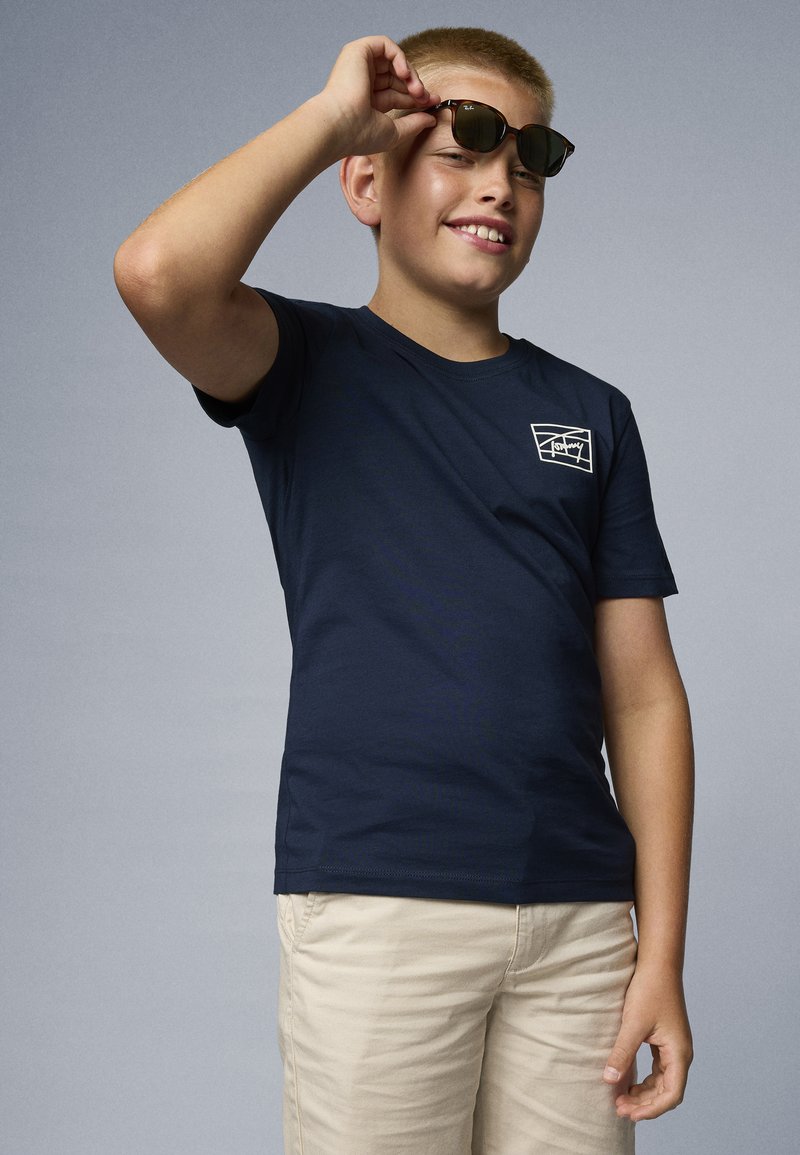 Marineblå bomull T-skjorte med korte ermer, med en hvit grafikk på brystet. Lys beige shorts; holder brune solbriller.