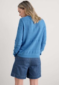 Zils adīts džemperis ar teksturētām dimantu rakstiem, ribotām piedurknēm un apakšmalu. Savienots ar džinsu šortiem ar vienkāršu dizainu.